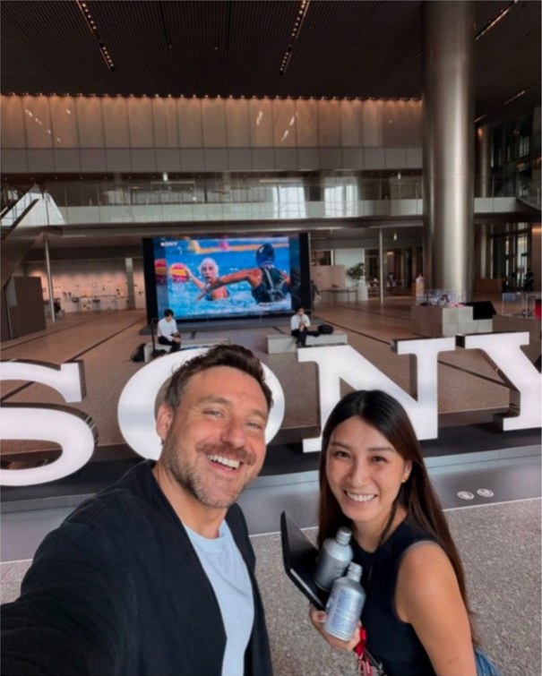 Two people take a selfie in front of the SONY letters in the lobby of Sony Group headquarters, with a video wall behind them