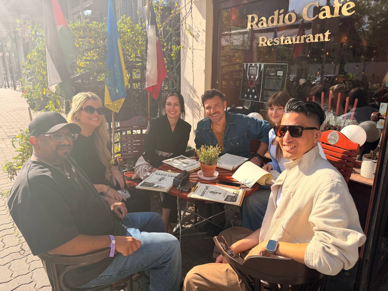 Six people around a café table in the sun, smiling at the camera