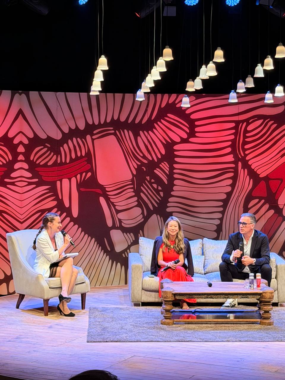 Three people in a panel discussion on a stage with a red patterned backdrop and hanging lamps