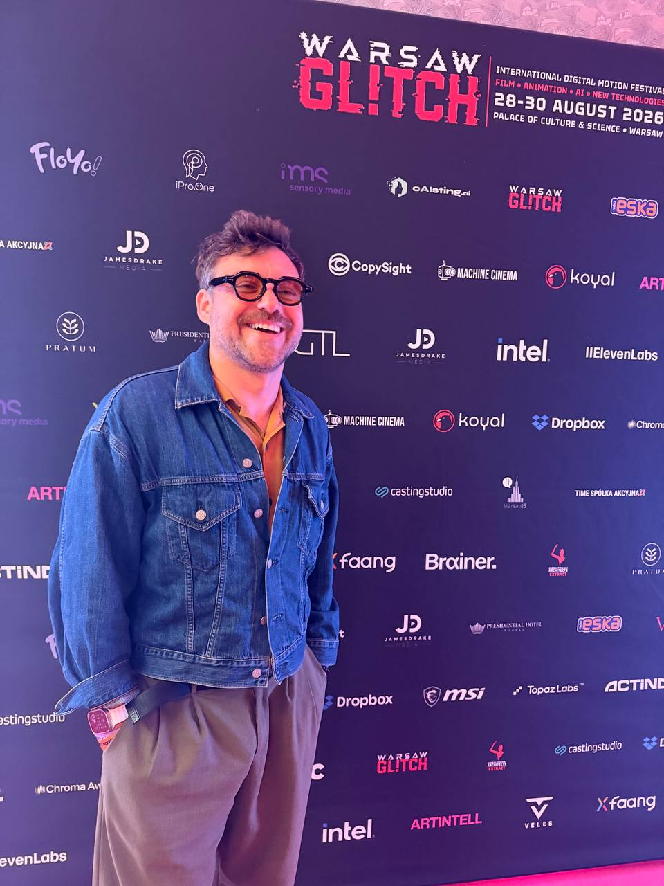 A man in a denim jacket at the Warsaw Glitch partner wall, with the festival dates and the CopySight logo among the sponsors